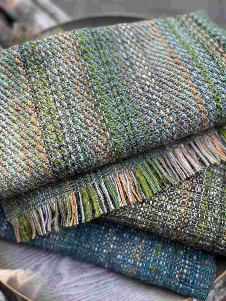 A close-up of three handwoven blankets showcasing intricate textures and colors in greens, blues, and earthy tones.
