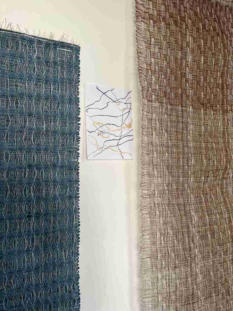 Textured woven artworks displayed on a wall, featuring a dark blue textile on the left, a light brown textile on the right, and a small abstract drawing in the center.