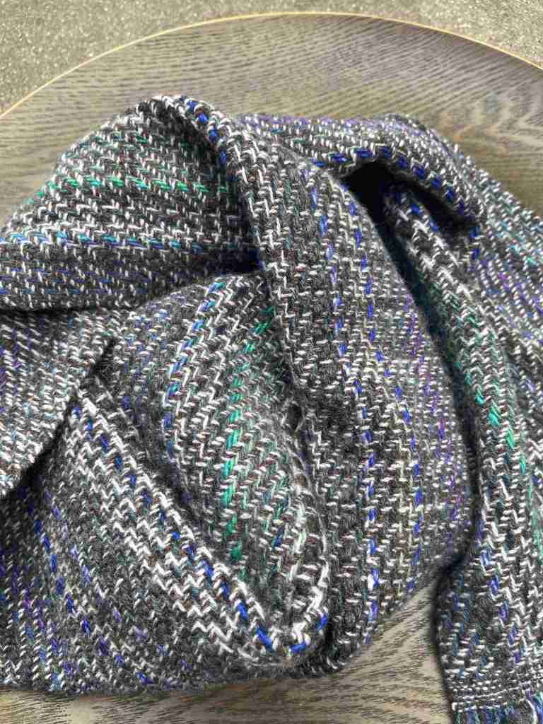 A close-up of a handwoven scarf displaying a mix of gray, purple, and green threads, elegantly folded on a wooden surface.