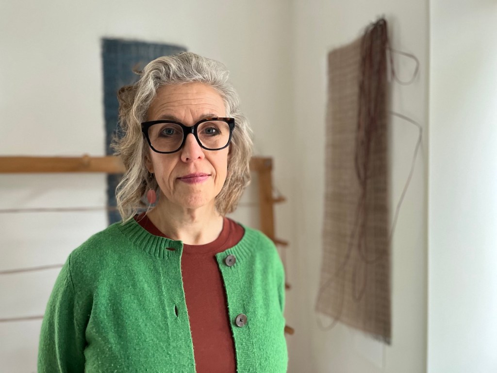 A woman with curly gray hair, wearing a green cardigan and a rust-colored top, stands in a studio with woven textiles hanging in the background.