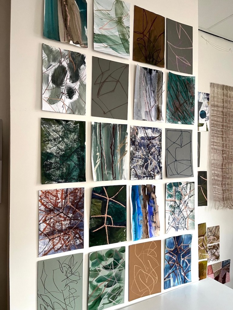 A wall display featuring a grid of various abstract paintings and mixed media artworks in different colors and textures.