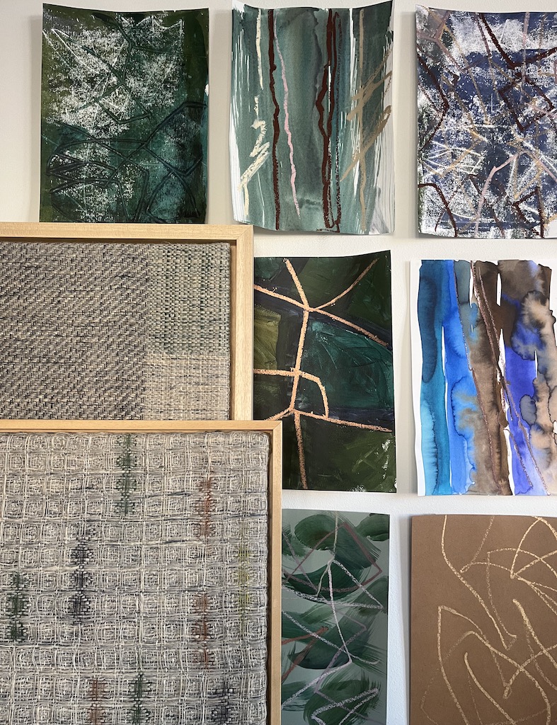 Collection of artworks and textile pieces displayed on a wall, featuring abstract paintings and woven fabric patterns.