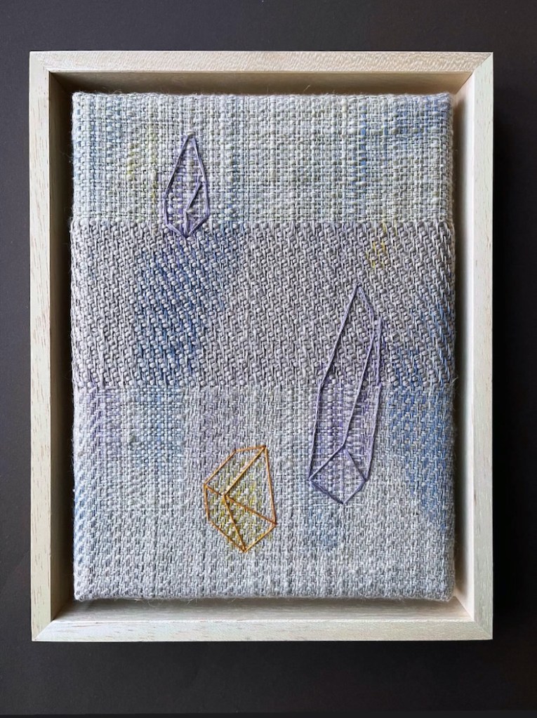 Textile artwork featuring geometric embroidered shapes on a textured background, framed in light wood.