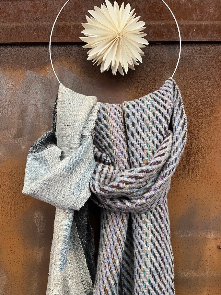 Two handwoven scarves displayed together, one in light grey tones and the other with a multicolored zigzag pattern, hanging from a circular decorative ring with a paper star ornament in the background.