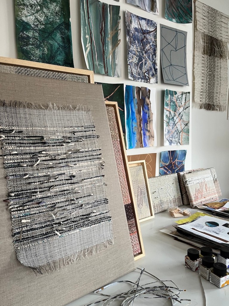 An art studio featuring woven textiles on display, with various pieces mounted on the wall and scattered artworks on a work table.