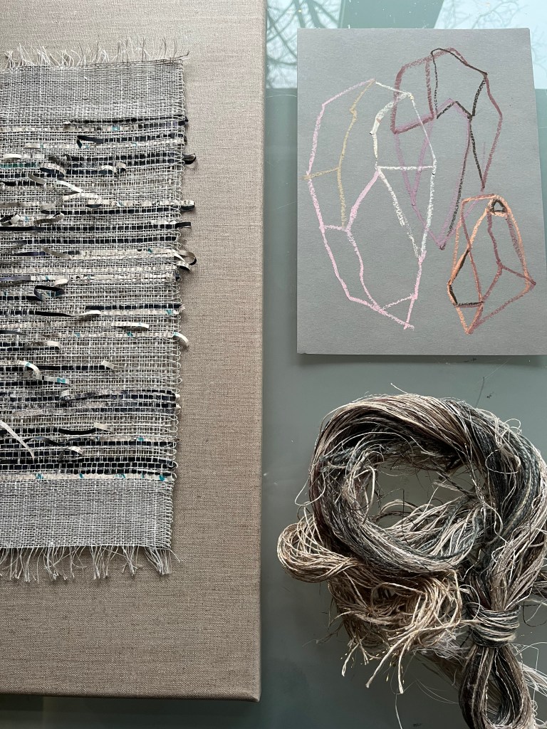 A textured woven fabric on the left, a grey card with abstract geometric shapes in pastel colours in the centre, and a bundle of natural fibres on the right, all displayed on a glass surface.