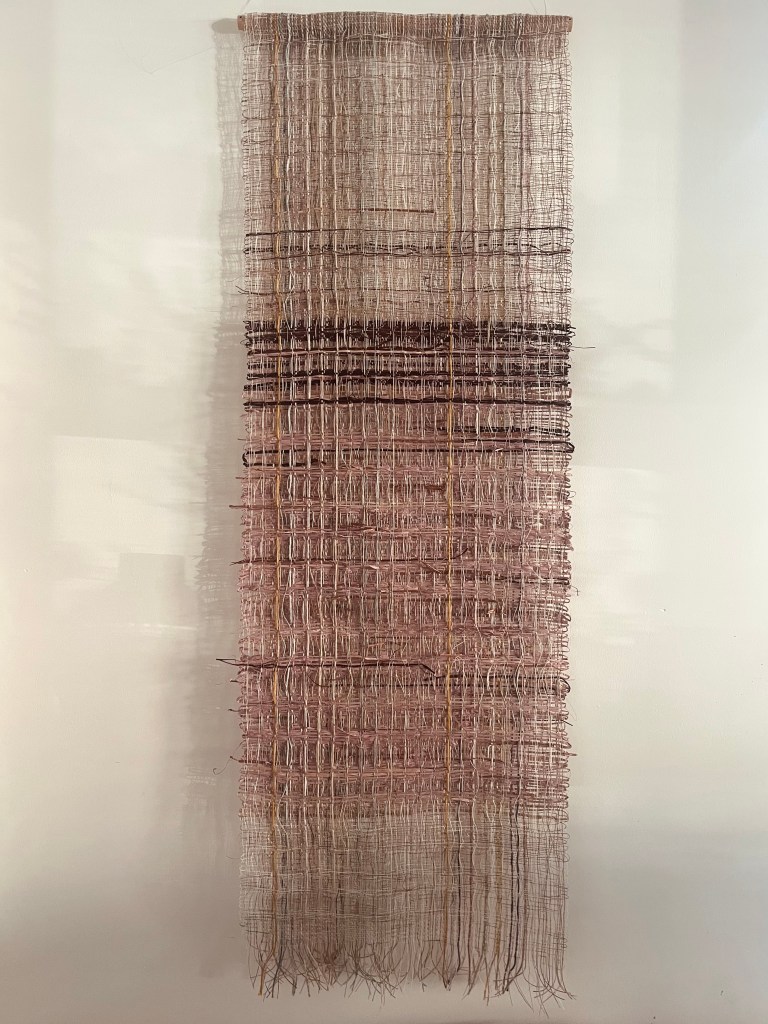 A vertical woven wall hanging featuring a textured pattern in shades of beige, brown, and pink, displayed against a white background.