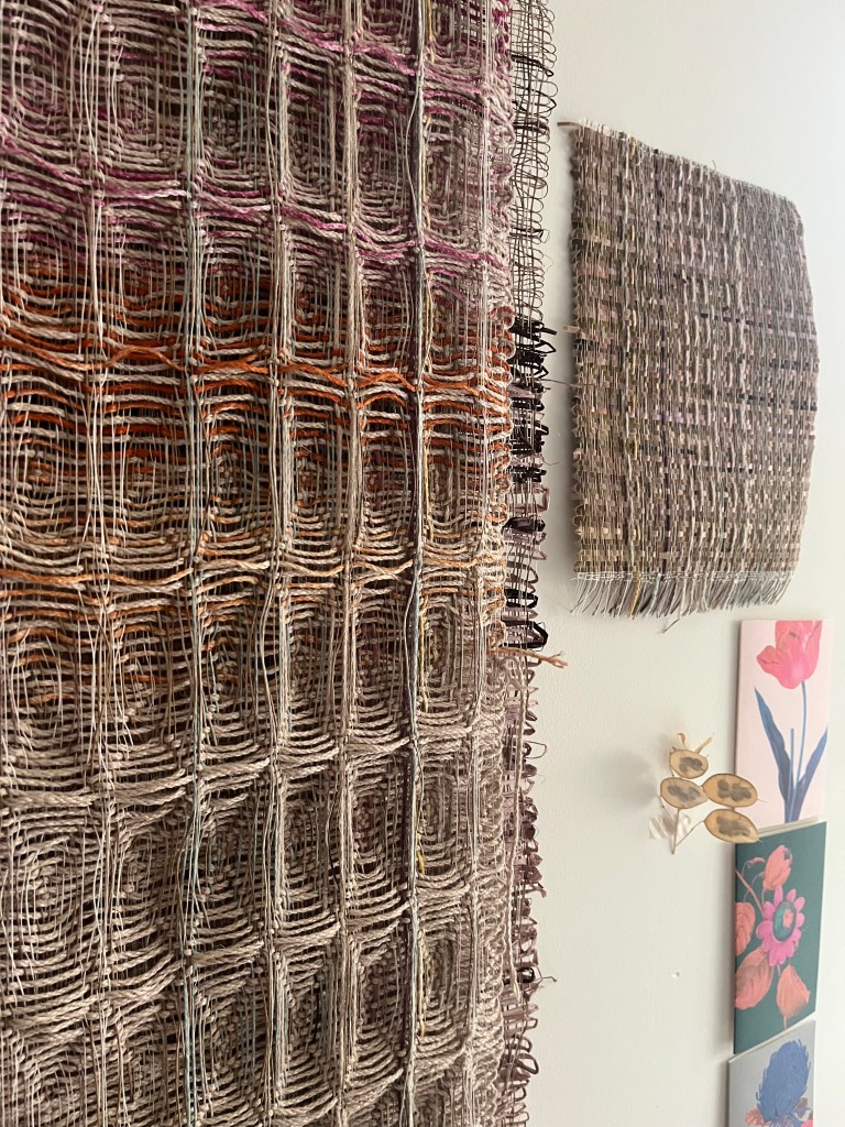 A wall displaying two woven textile pieces, one large with a grid pattern in earthy tones, and another smaller, textured piece above it. Nearby, several floral artworks are mounted on the wall.